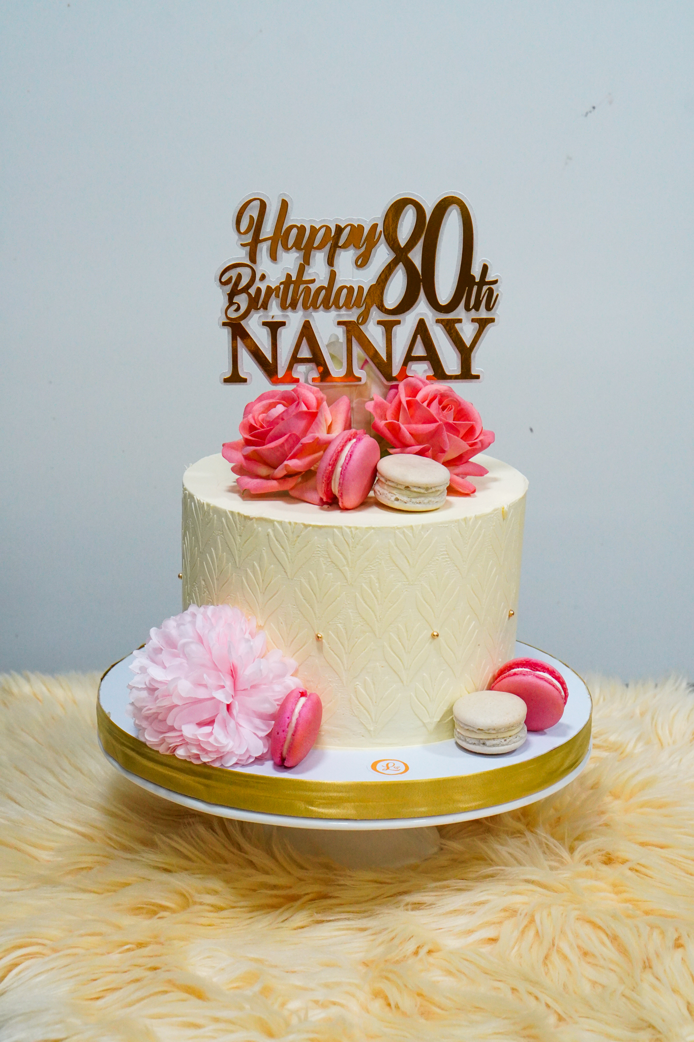 Elegant cream birthday cake with pink flowers, macarons, and a gold topper for an 80th birthday