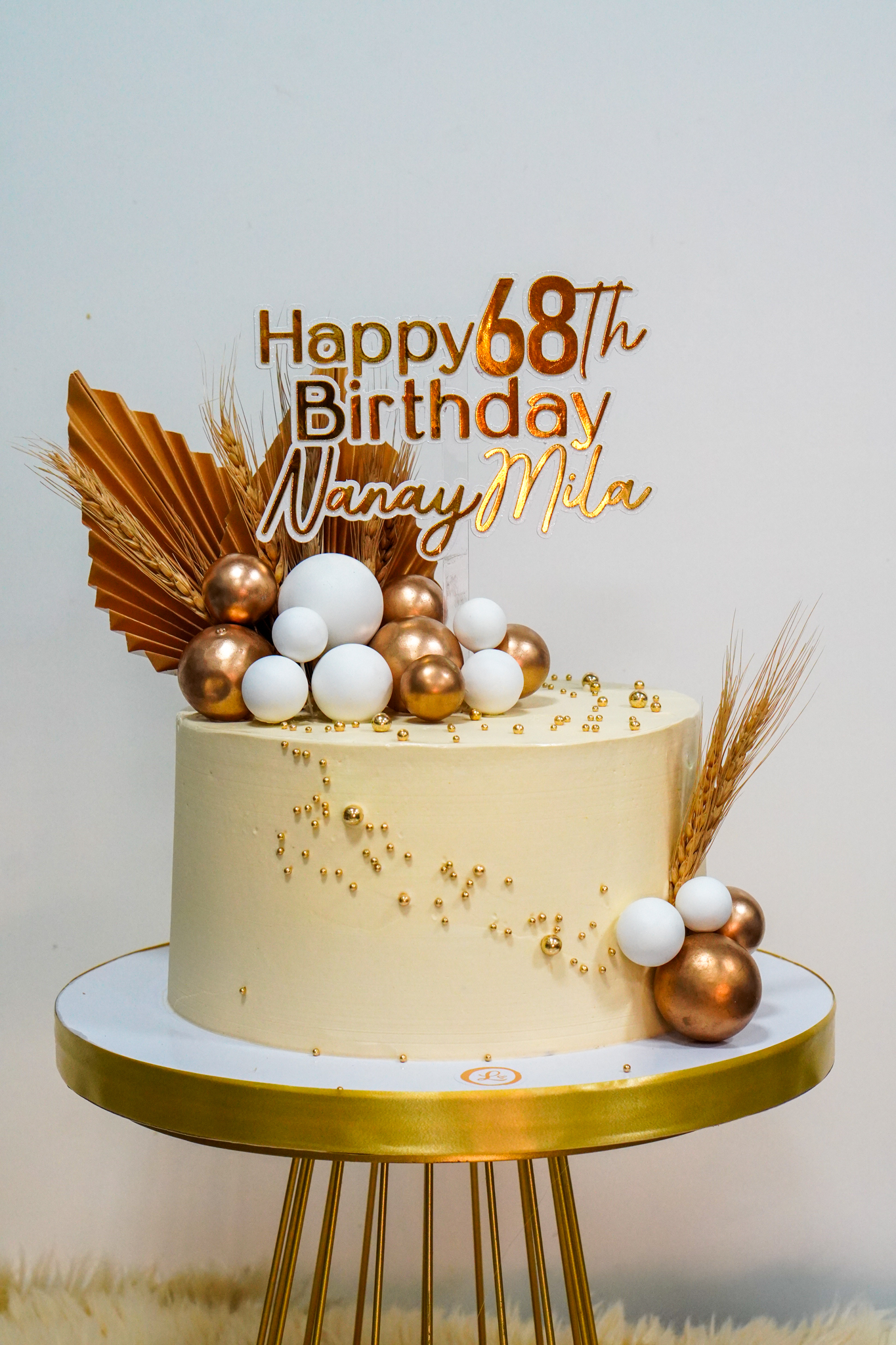 Elegant cream birthday cake with gold and white balloon clusters, dried leaves, and wheat stalk accents
