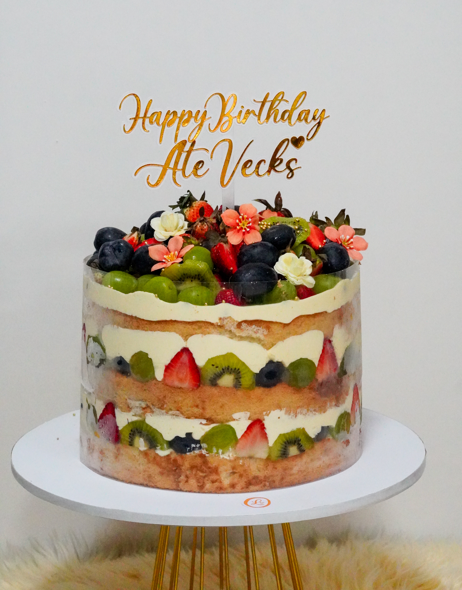 Naked-style birthday cake with alternating layers of sponge cake, cream, and fresh fruits including strawberries, kiwis, blueberries, grapes, and topped with assorted fruits and edible flowers, with a 'Happy Birthday Ate Vecks' gold topper