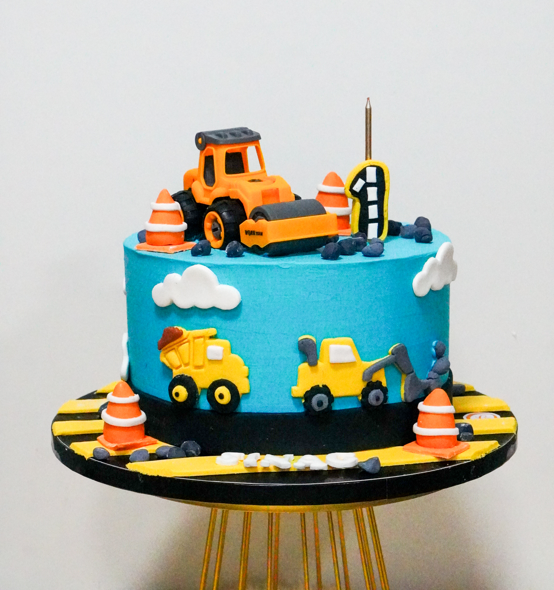 Blue buttercream birthday cake with construction vehicle toppers, traffic cones, clouds, and road details, featuring a number 1 candle