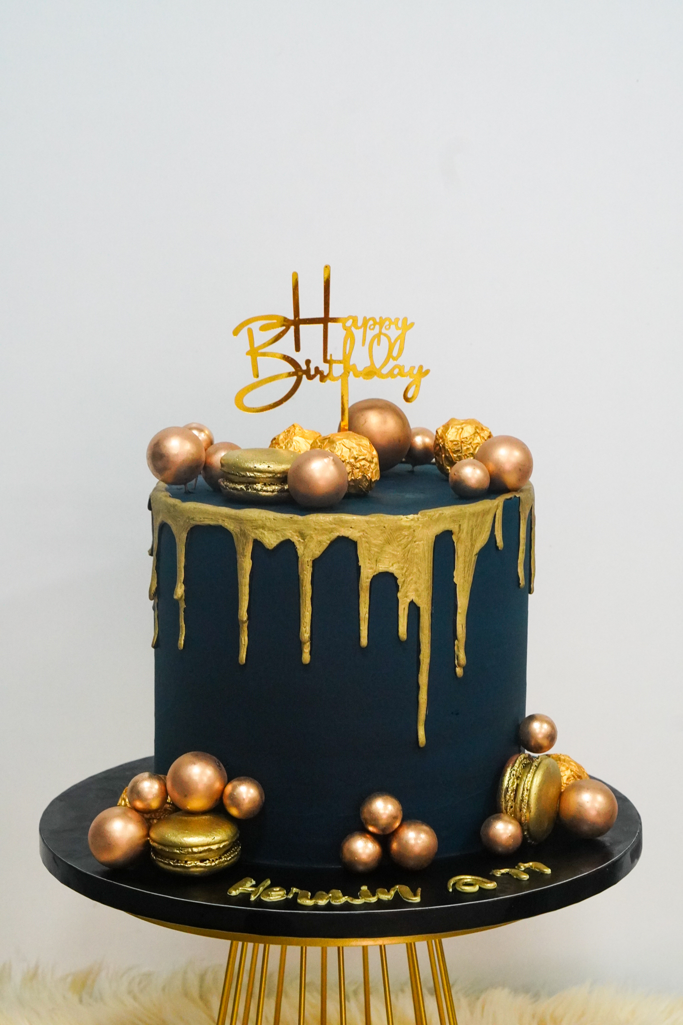 Black buttercream birthday cake with gold drip design, decorated with gold and bronze spheres, macarons, and Ferrero Rocher chocolates, topped with a gold 'Happy Birthday' topper
