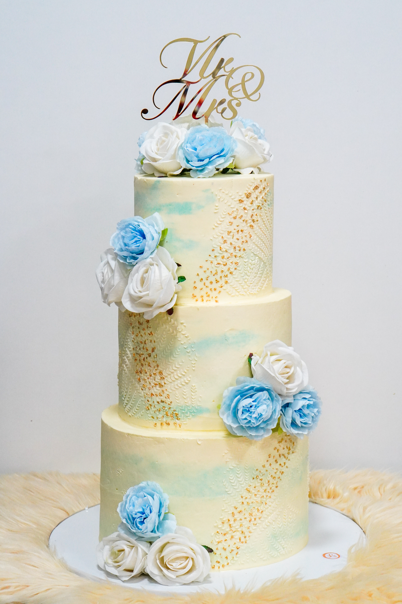 Three-tier buttercream wedding cake with textured gold accents, light blue watercolor details, and decorated with white and light blue roses, topped with a gold 'Mr & Mrs' topper