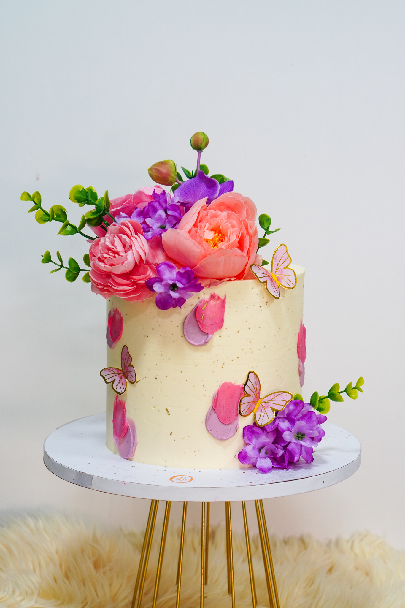 Single-tier buttercream cake decorated with large pink and purple flowers, green foliage, pink and purple textured patches, and gold-edged butterfly toppers