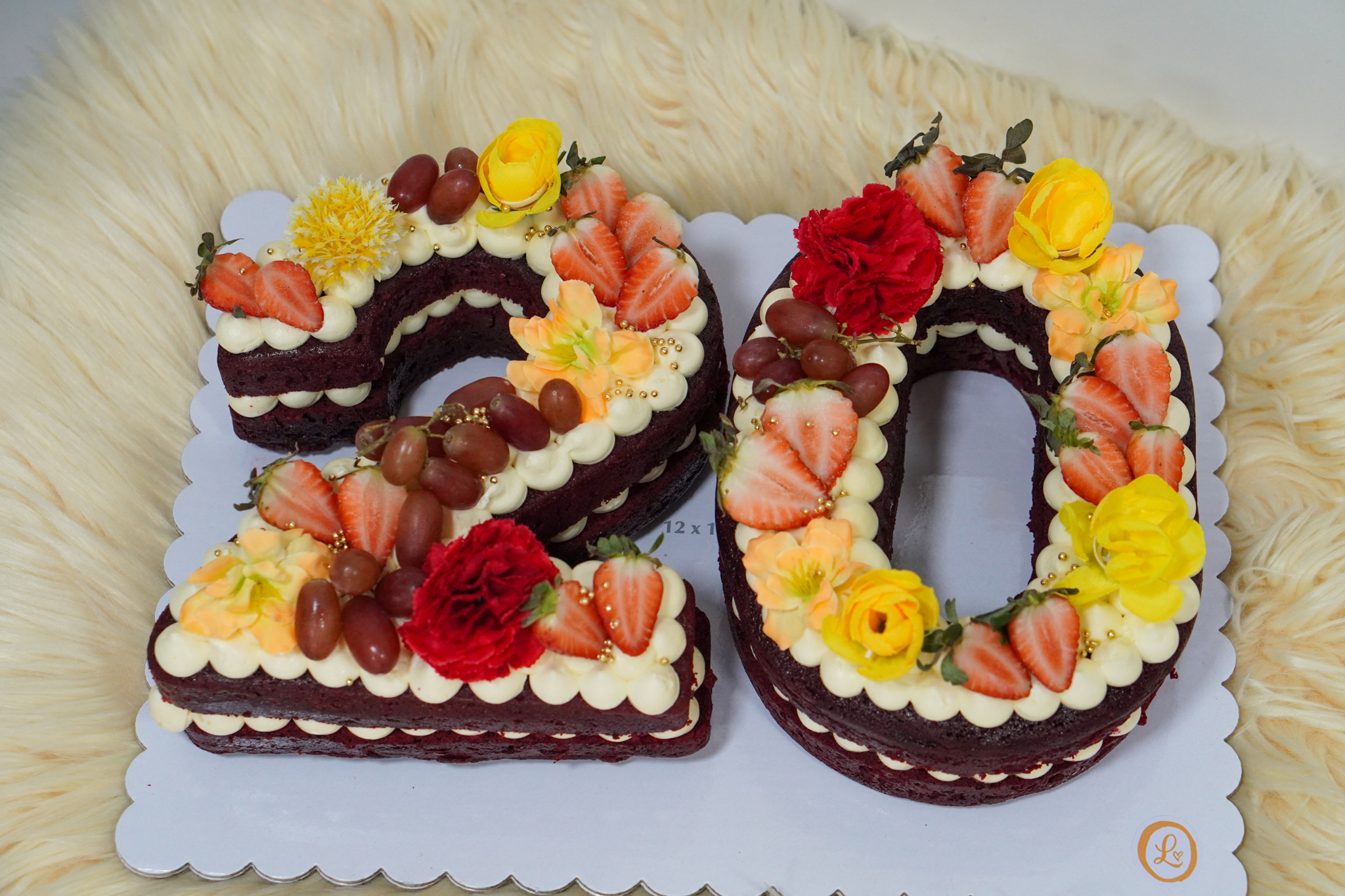 Number-shaped '20' cake with layers of red velvet sponge and cream, topped with strawberries, grapes, yellow and red flowers, and gold sprinkles