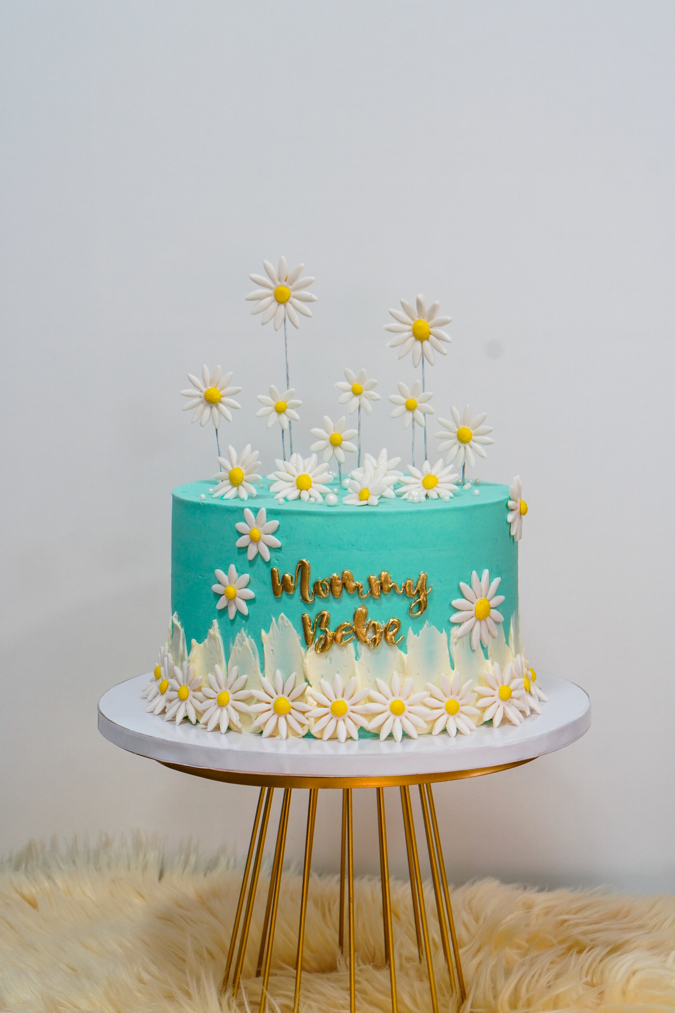 Teal buttercream cake with gold text 'Mommy Bebe', decorated with white and yellow daisy flowers