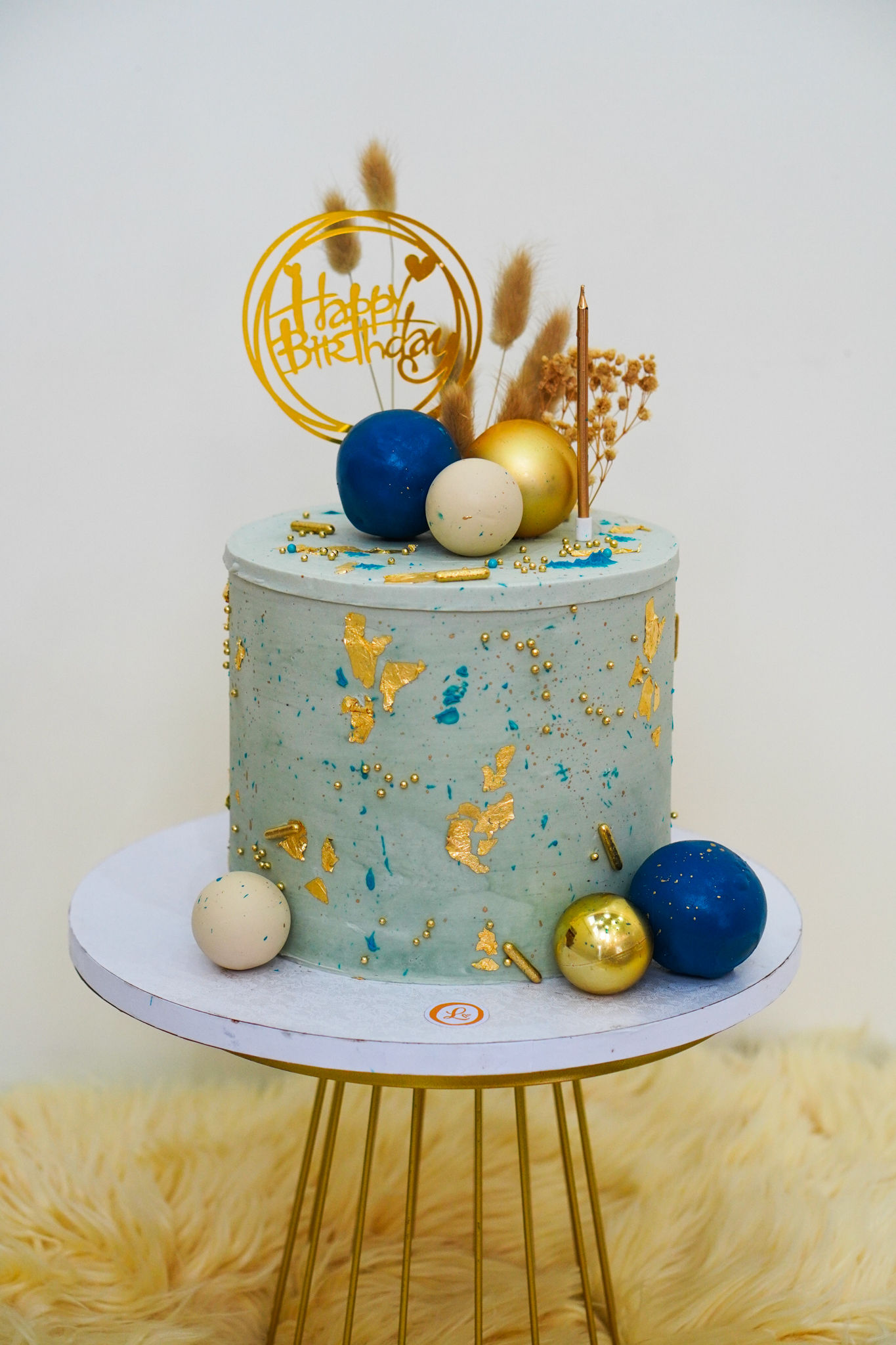 Blue buttercream birthday cake with gold leaf accents, spherical decorations in blue, gold, and cream, dried floral toppers, and a gold 'Happy Birthday' topper