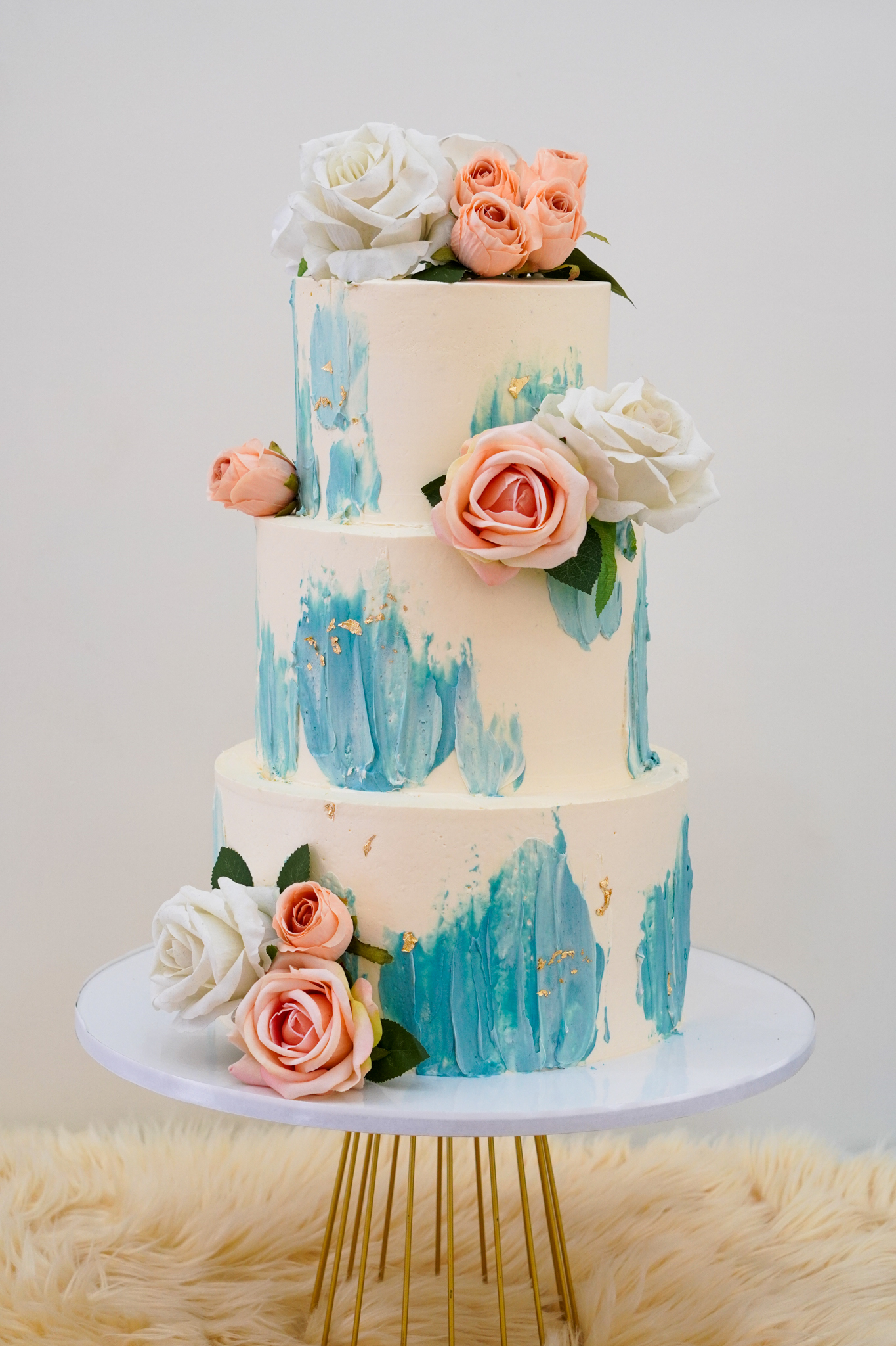 Three-tier cream buttercream cake with blue brushstroke design, gold leaf accents, and decorated with white and peach roses