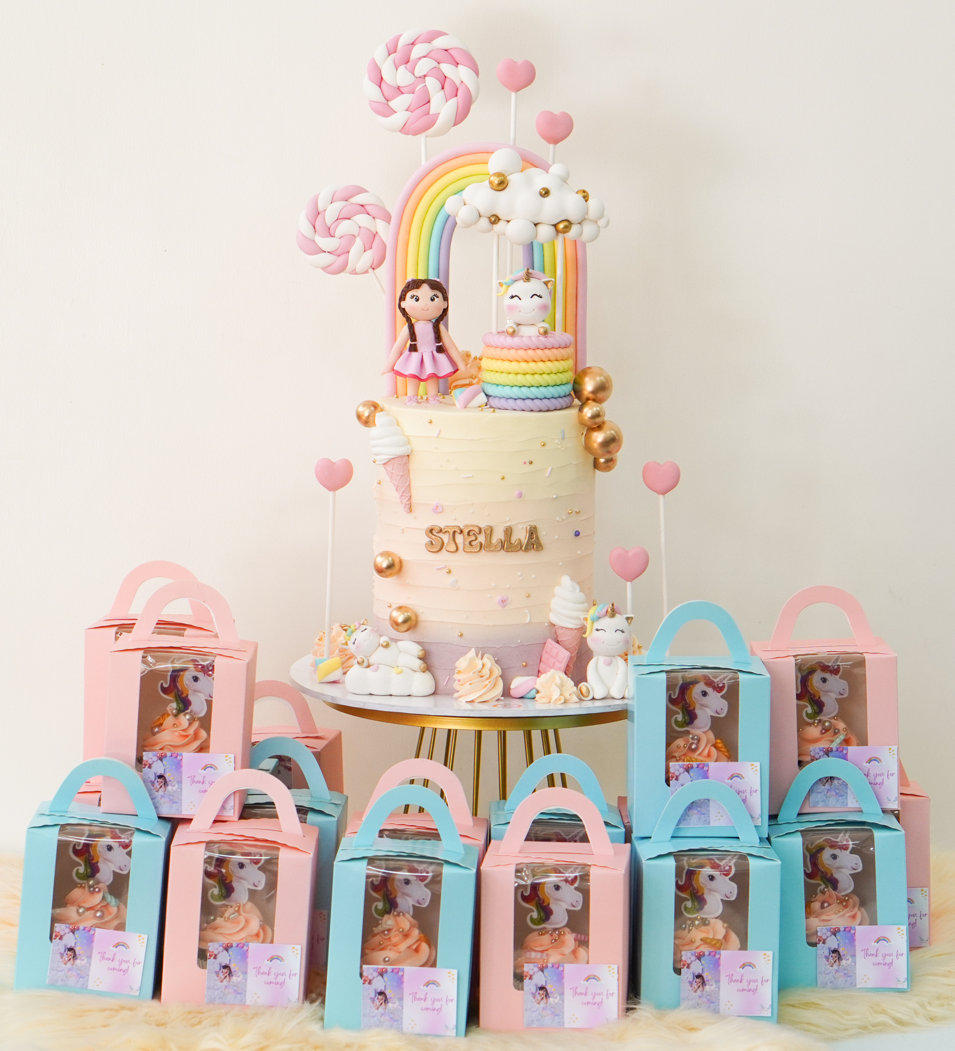 Pastel buttercream birthday cake with rainbow topper, doll figurine, unicorn figurine, ice cream cone decorations, and the name 'Stella', surrounded by pastel pink and blue favor boxes