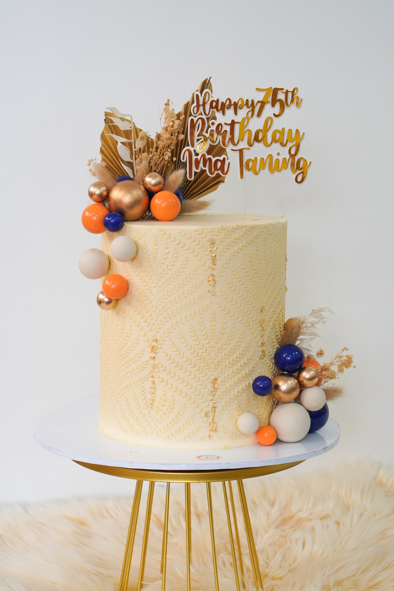 Elegant cream buttercream birthday cake with textured leaf pattern, adorned with gold, blue, orange, and white decorative spheres, dried florals, and a gold topper reading 'Happy 75th Birthday Ima Tansing'