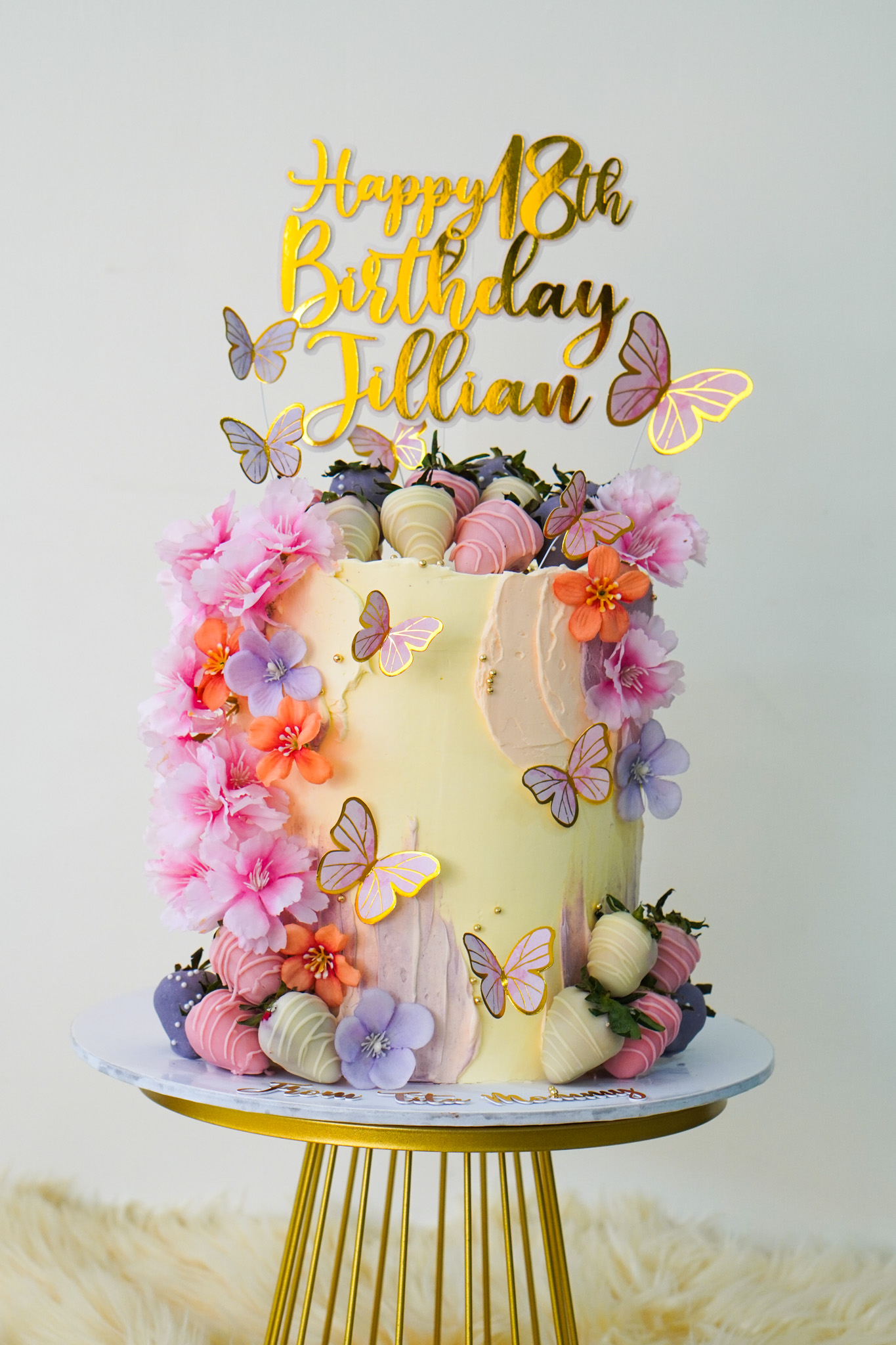 Pastel buttercream birthday cake with gold 'Happy 18th Birthday Jillian' topper, decorated with pink, purple, and orange flowers, butterflies, and chocolate-covered strawberries